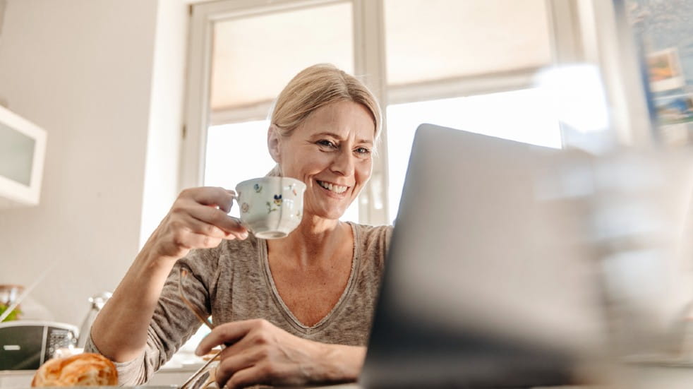 An older woman is sat looking at silver-coloured laptop. She holds a teacup in her right hand, she is sat in a light, bright room. 